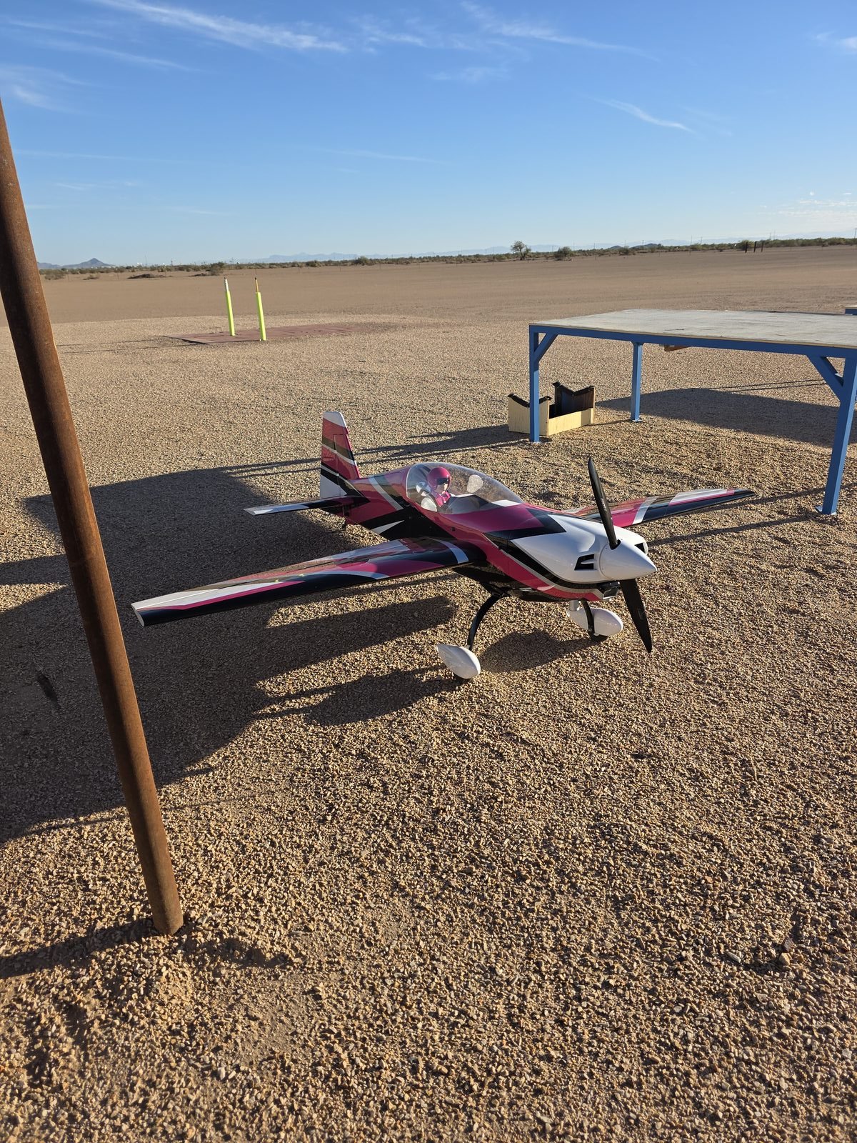 Pink, black, and white RC airplane on gravel near the sun shade support pole at the club field